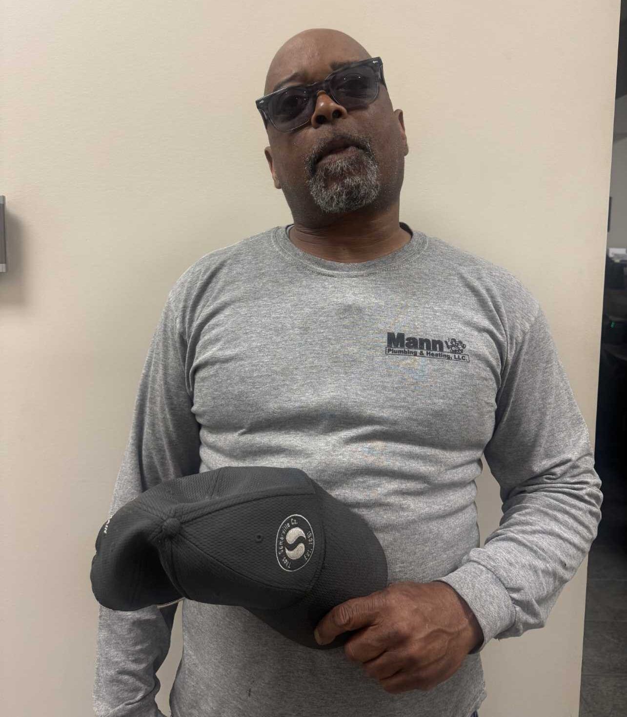 Man wearing sunglasses and a gray long-sleeved shirt holding a cap, standing in a room.