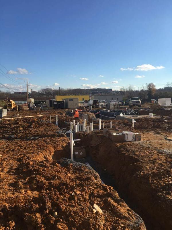 Construction site with trenches, pipes, and a worker in an orange vest under a blue sky.