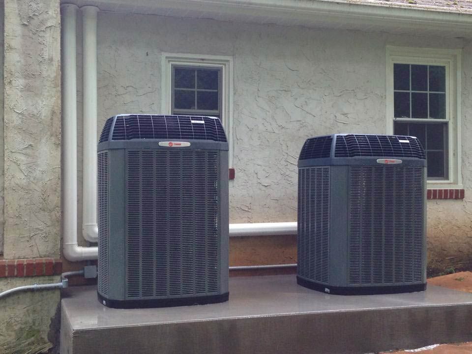 Two gray air conditioning units outside a building with two windows.