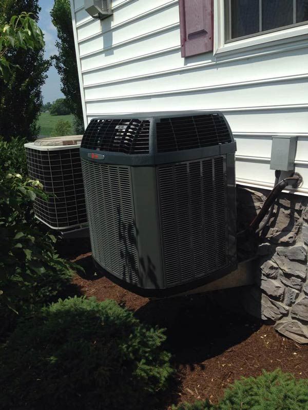 Two air conditioning units beside a white house with brown mulch and greenery.