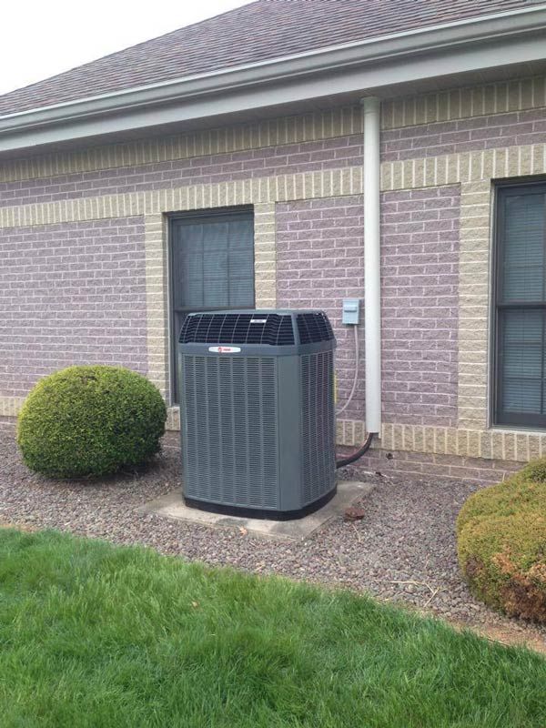 Air conditioning unit next to a brick building and shrubbery, on gravel.