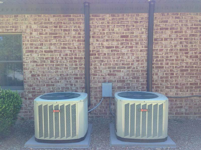 Two air conditioning units against a brick wall, with vertical pipes and an electrical box.