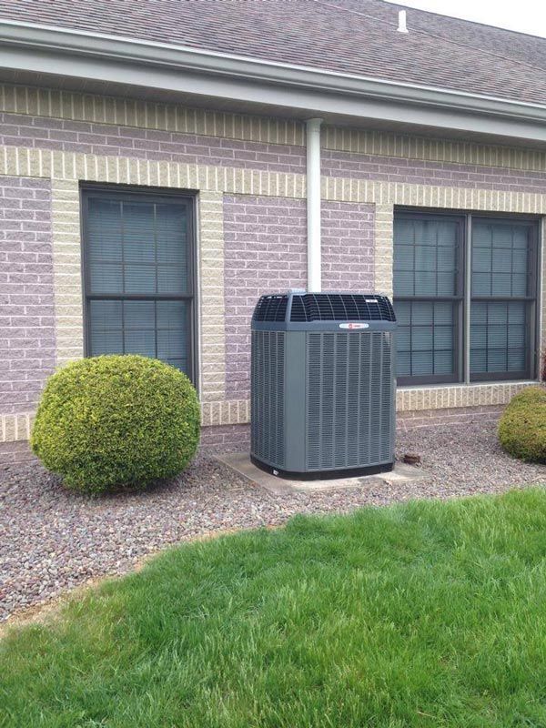 Gray HVAC unit next to a building with brick exterior and two windows. Green bushes and grass.