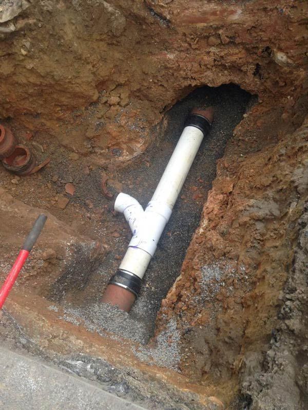Pipes in a trench. White and brown pipes joined by a Y-shaped fitting. The trench is in dirt with gray gravel.