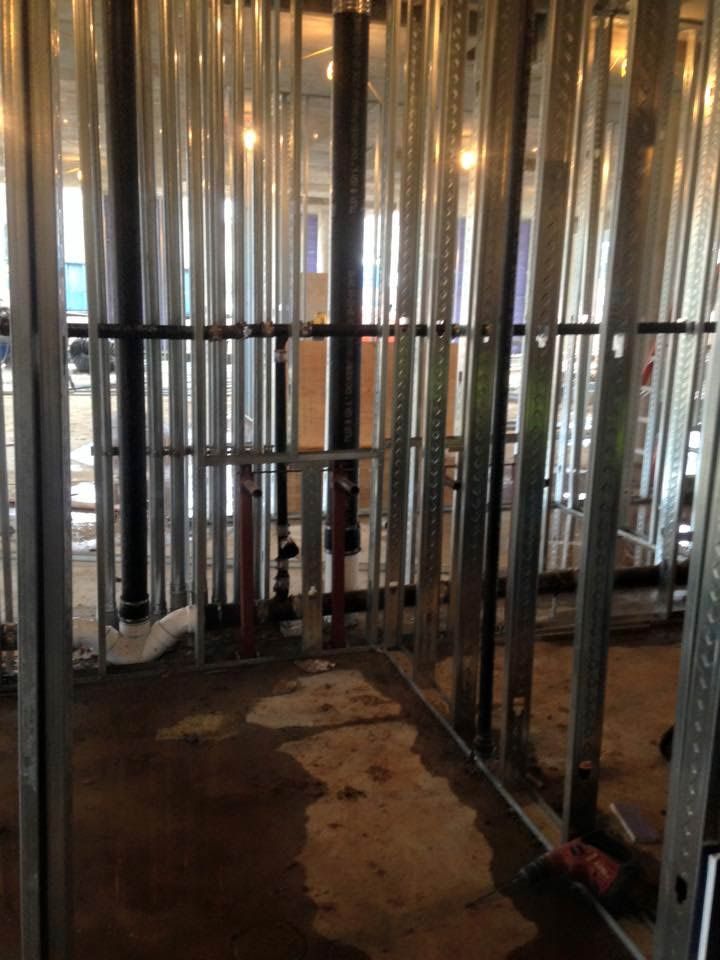 Metal studs and plumbing in a room under construction. Gray and black pipes run vertically and horizontally. Brown floor.