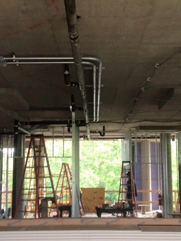 Construction site interior with pipes and electrical conduit on ceiling; ladders and view of trees visible.