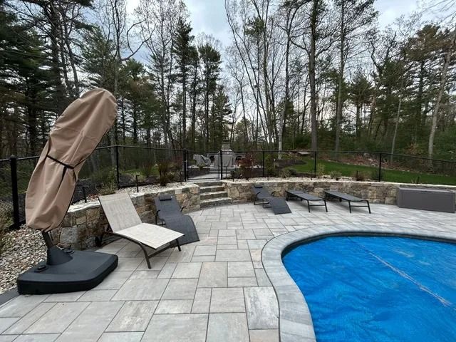 Patio with blue pool, lounge chairs, fire pit, and stone seating area surrounded by trees