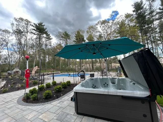 Outdoor hot tub with teal umbrella beside a pool, landscaped patio, and trees beyond
