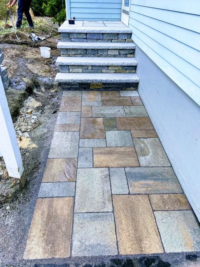 Stone paver walkway leading to front steps beside a light blue house and rocky landscape