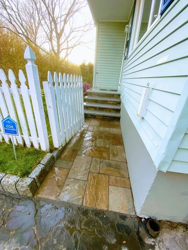 Narrow side walkway with stone pavers beside a white house and picket fence leading to steps.