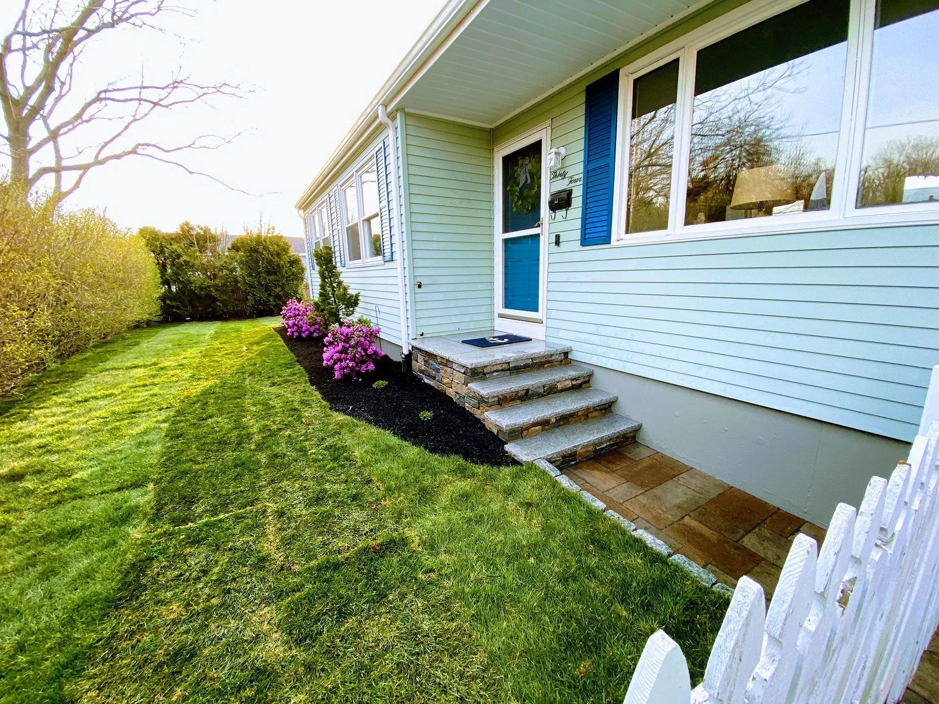 Blue house entrance with steps, white fence, and pink flowers beside a green lawn
