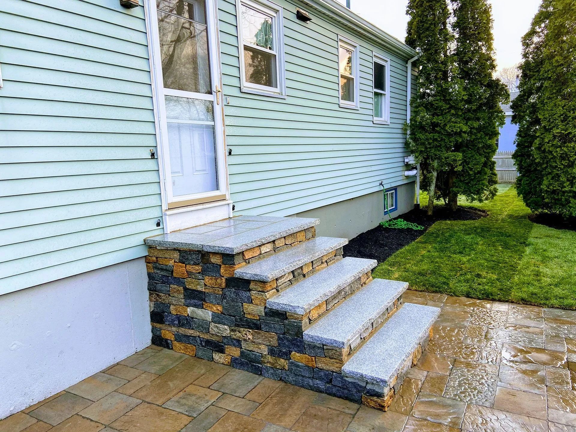 House side entrance with stone steps, white door, and light blue siding beside a wet patio and lawn