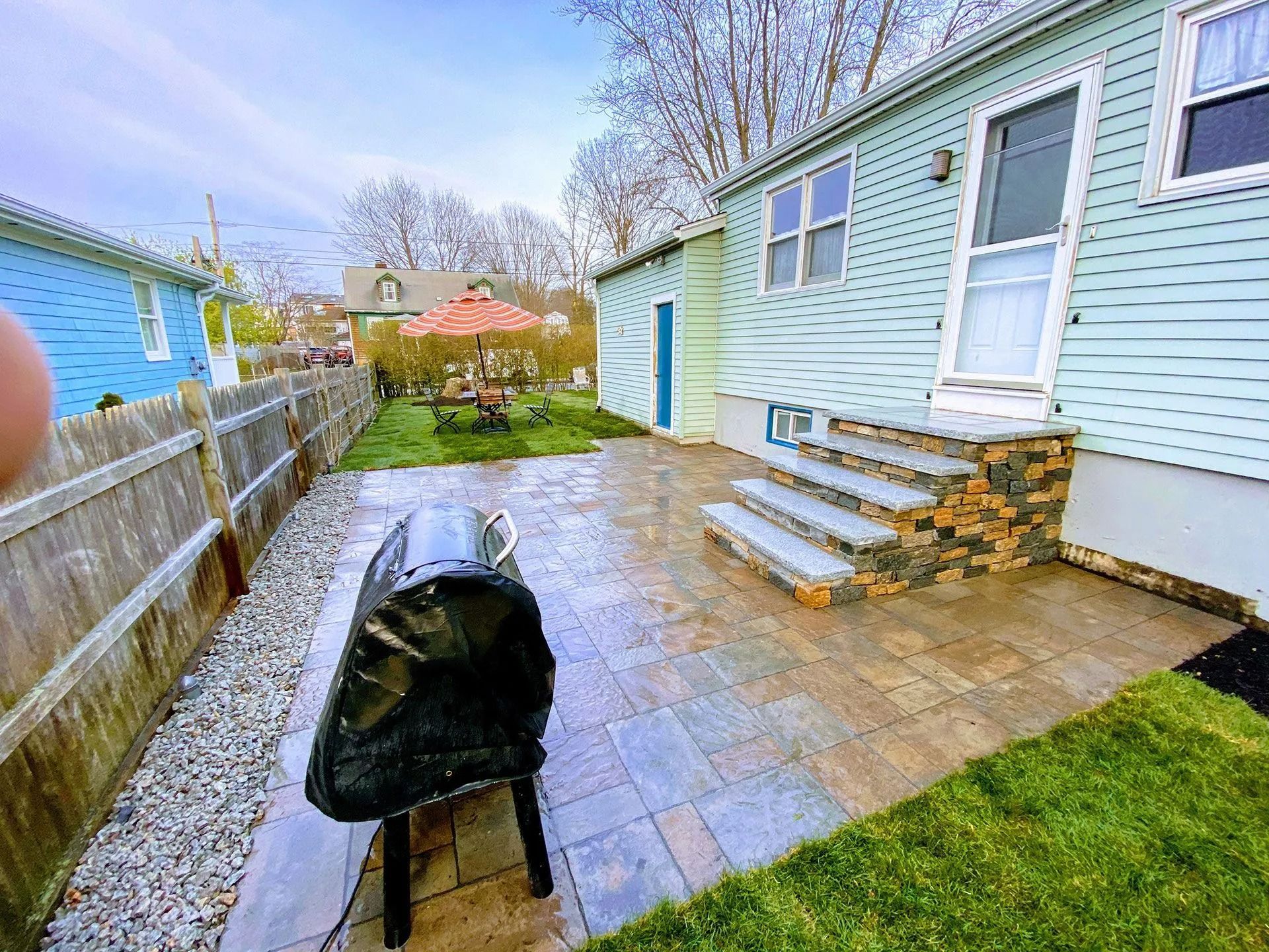 Backyard patio with a covered grill, steps, and a light blue house with a fenced yard.