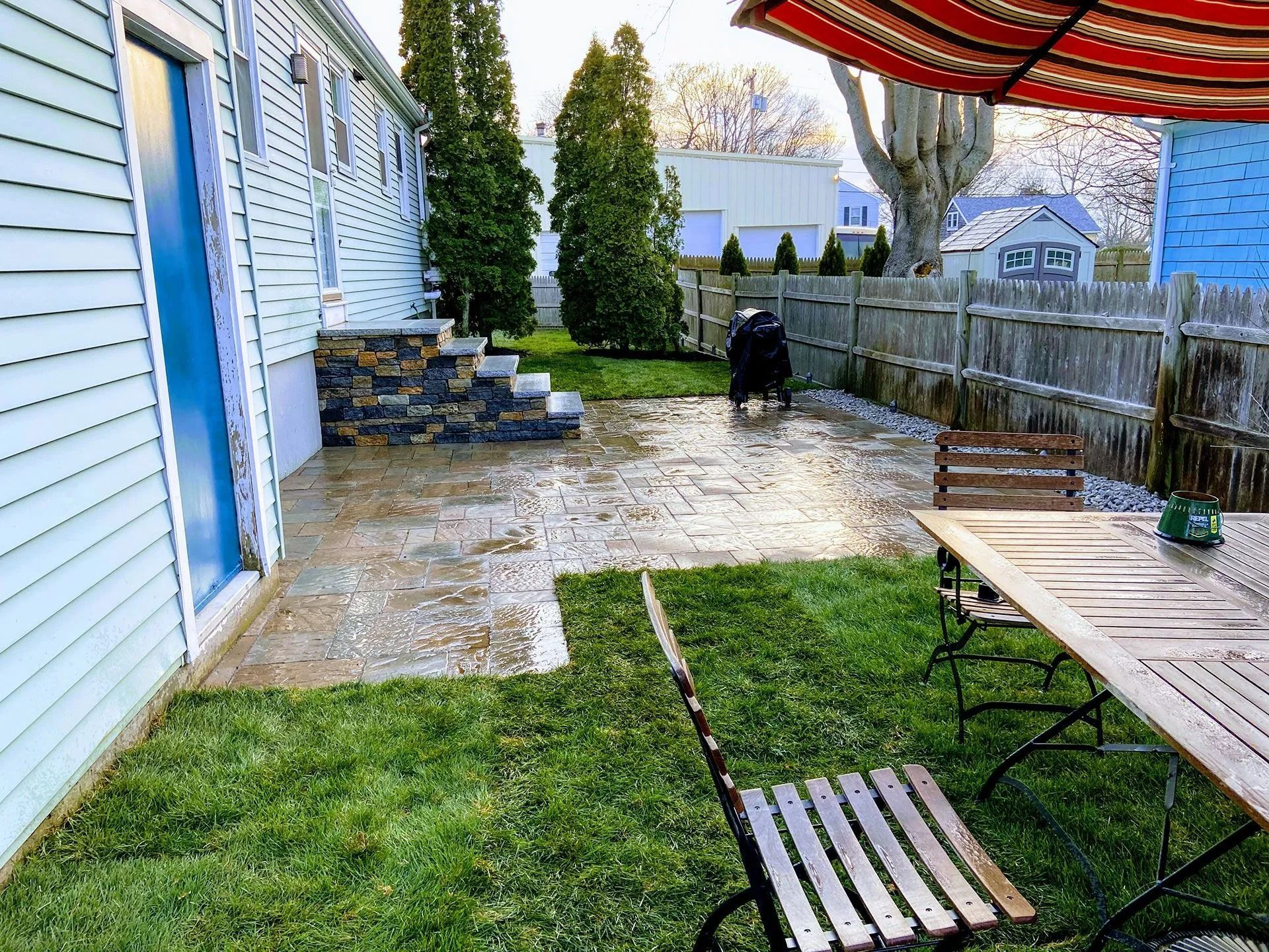 Backyard patio with wet stone, green lawn, wooden table, chairs, and blue house siding.