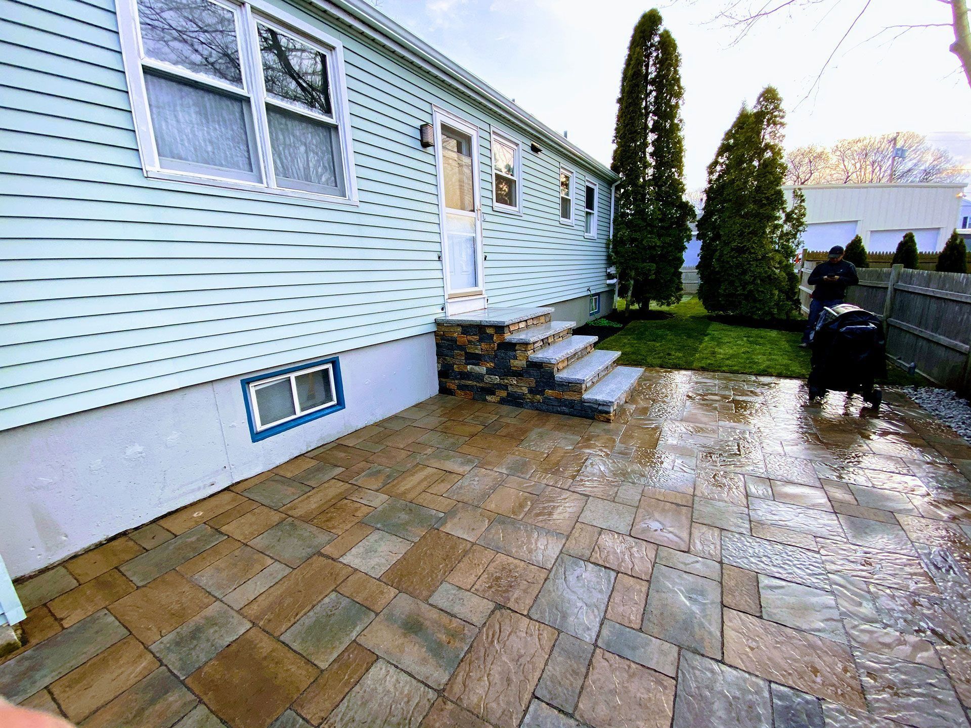 Back patio with wet stone pavers beside a white house, steps, shrubs, and a covered grill.