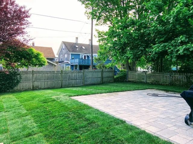Backyard with green lawn, patio, fence, trees, and houses beyond the yard