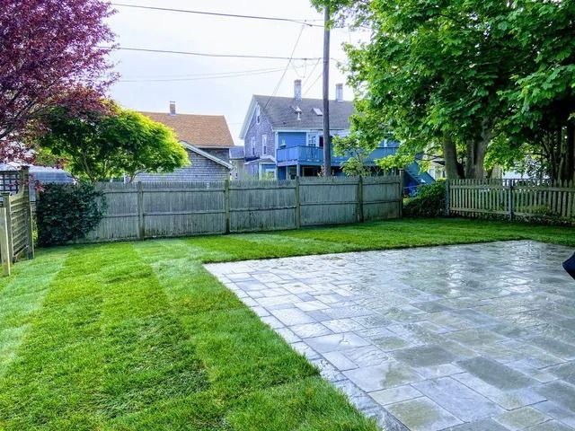 Backyard with fresh green lawn, paved patio, wooden fence, and houses beyond the trees