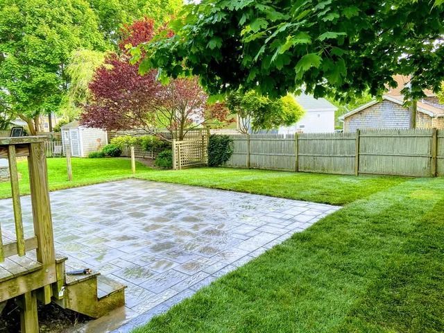 Backyard patio with gray pavers, green lawn, wooden fence, and trees