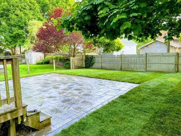 Backyard patio with gray pavers, green lawn, wooden deck steps, and trees along a fence