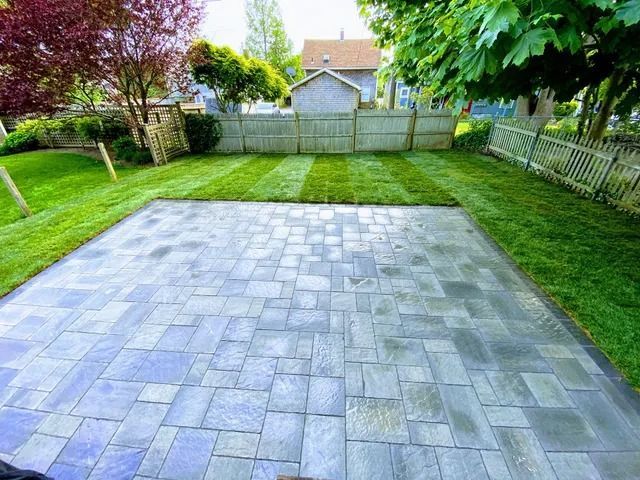 Backyard with a paved patio, green lawn, and fence under trees