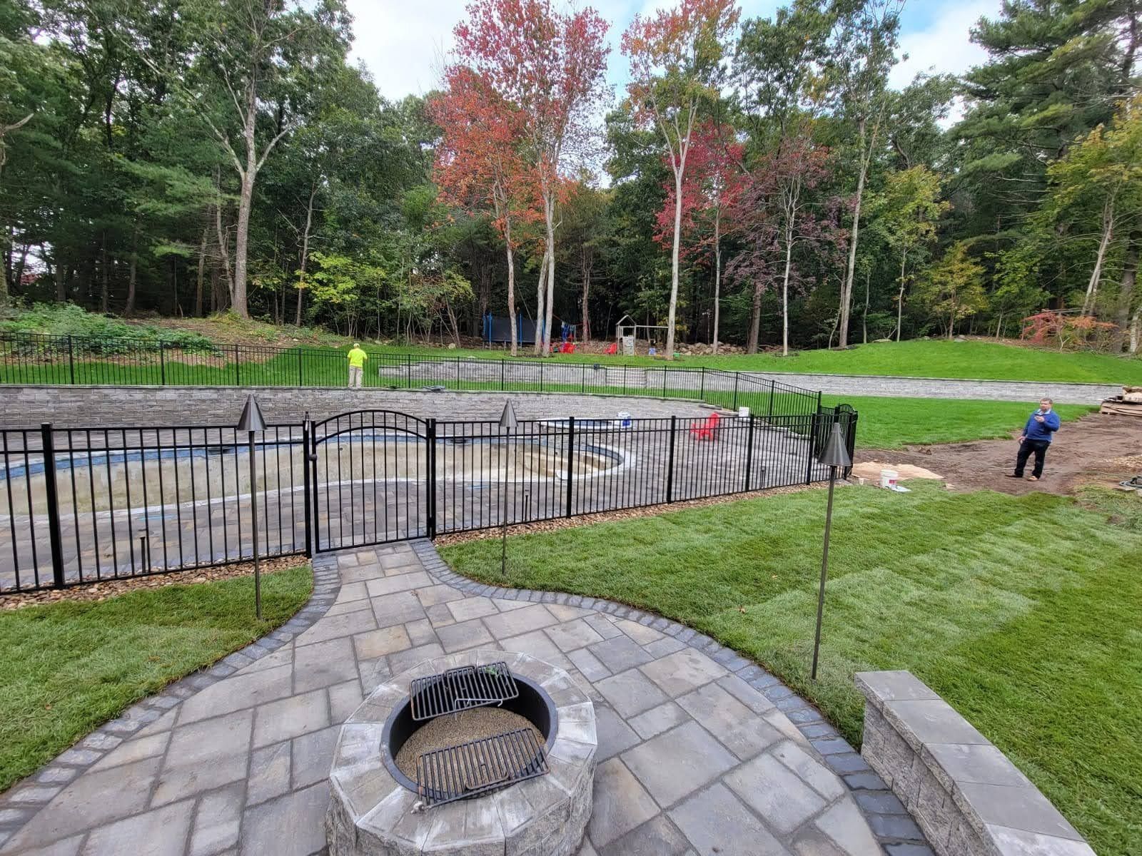 Park with fenced pond, stone patio, grassy lawn, and two people near the water under autumn trees