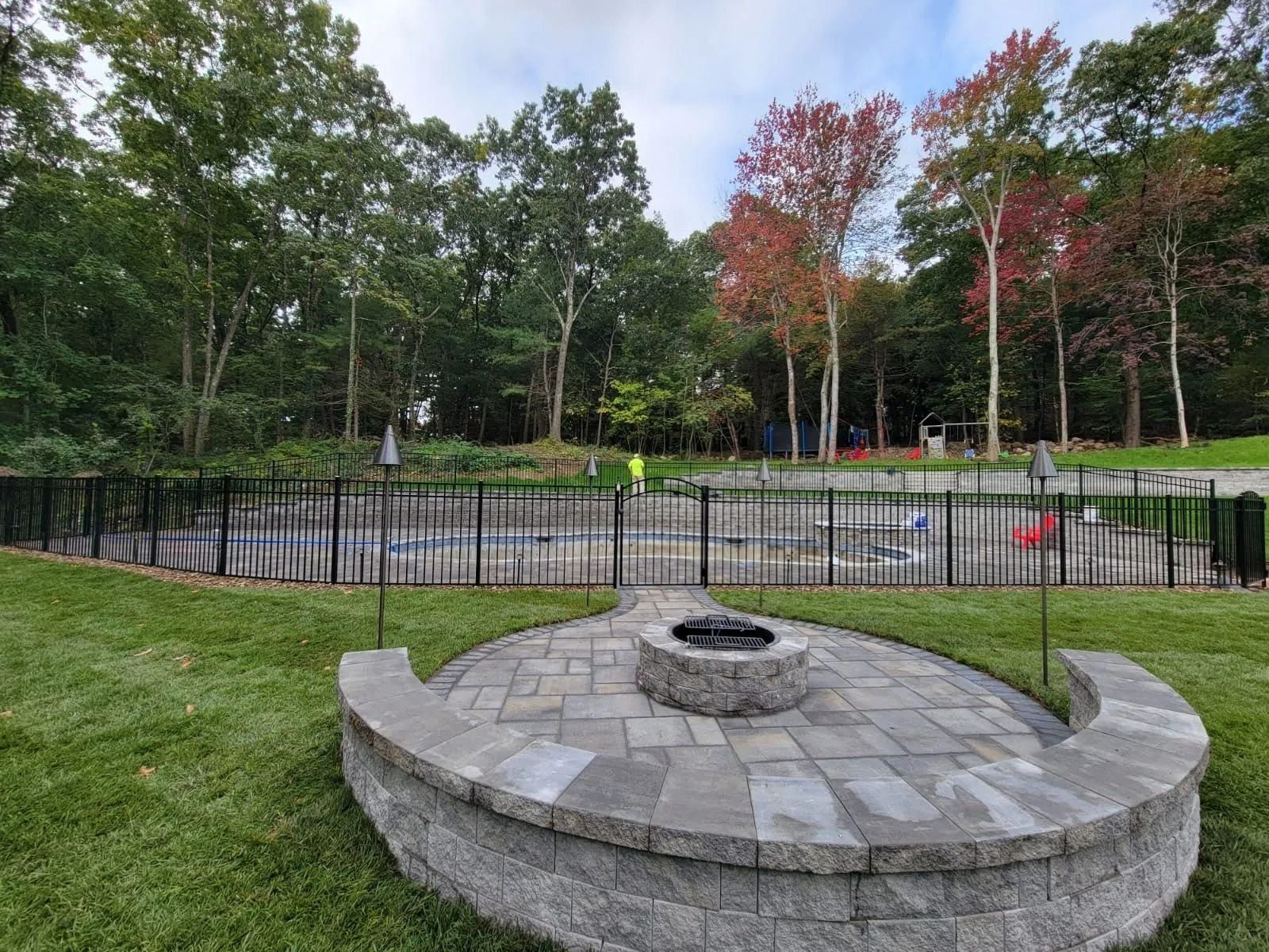 Circular stone fire pit by a fenced pool in a green wooded park