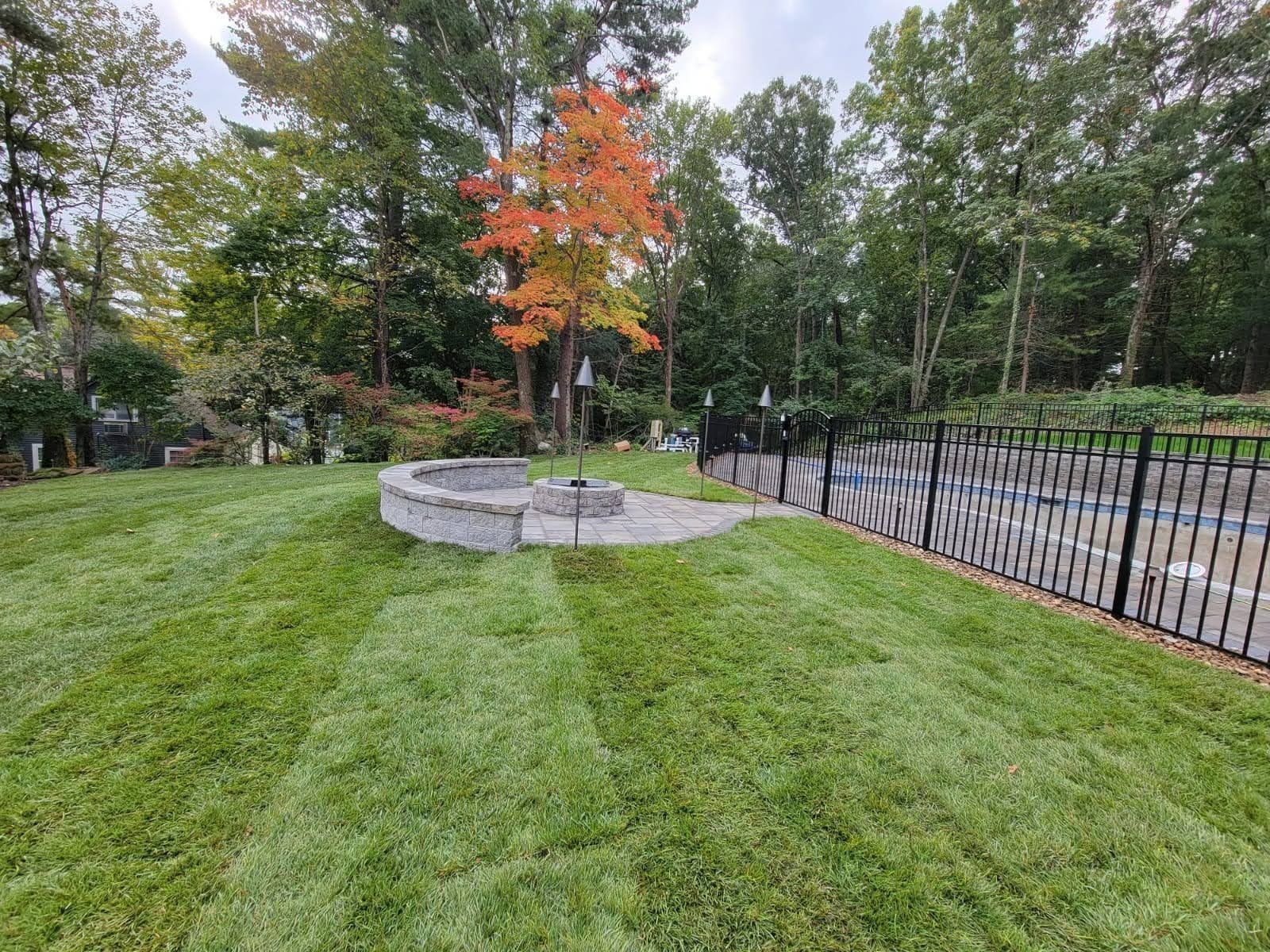 Grassy park with a small stone fire pit, a red-orange tree, and a black fence along the path.