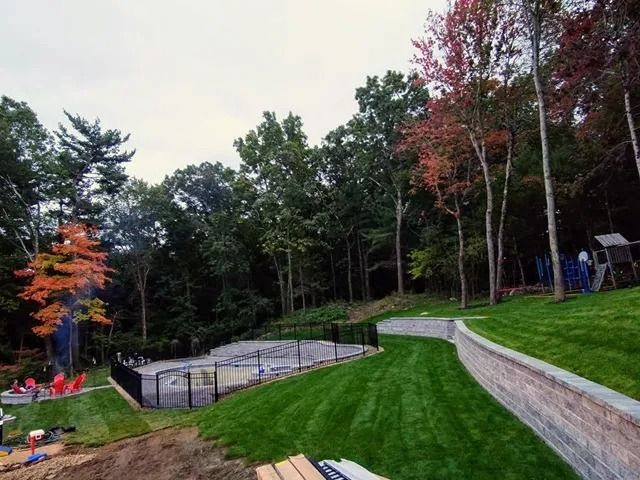 Sloped backyard with green lawn, stone retaining wall, trees, and a fenced in-ground pool