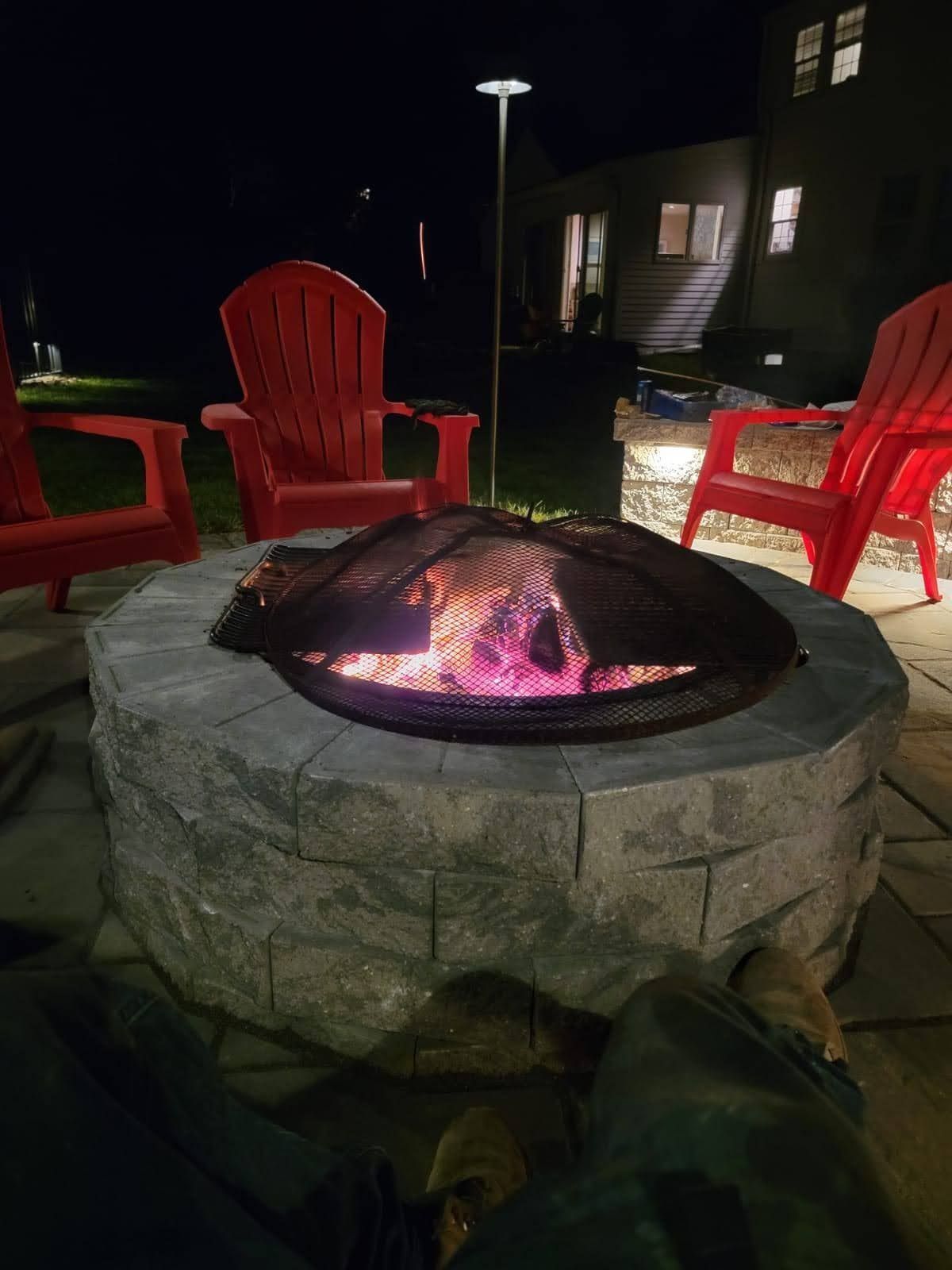 Stone fire pit glowing at night, surrounded by red Adirondack chairs in a backyard