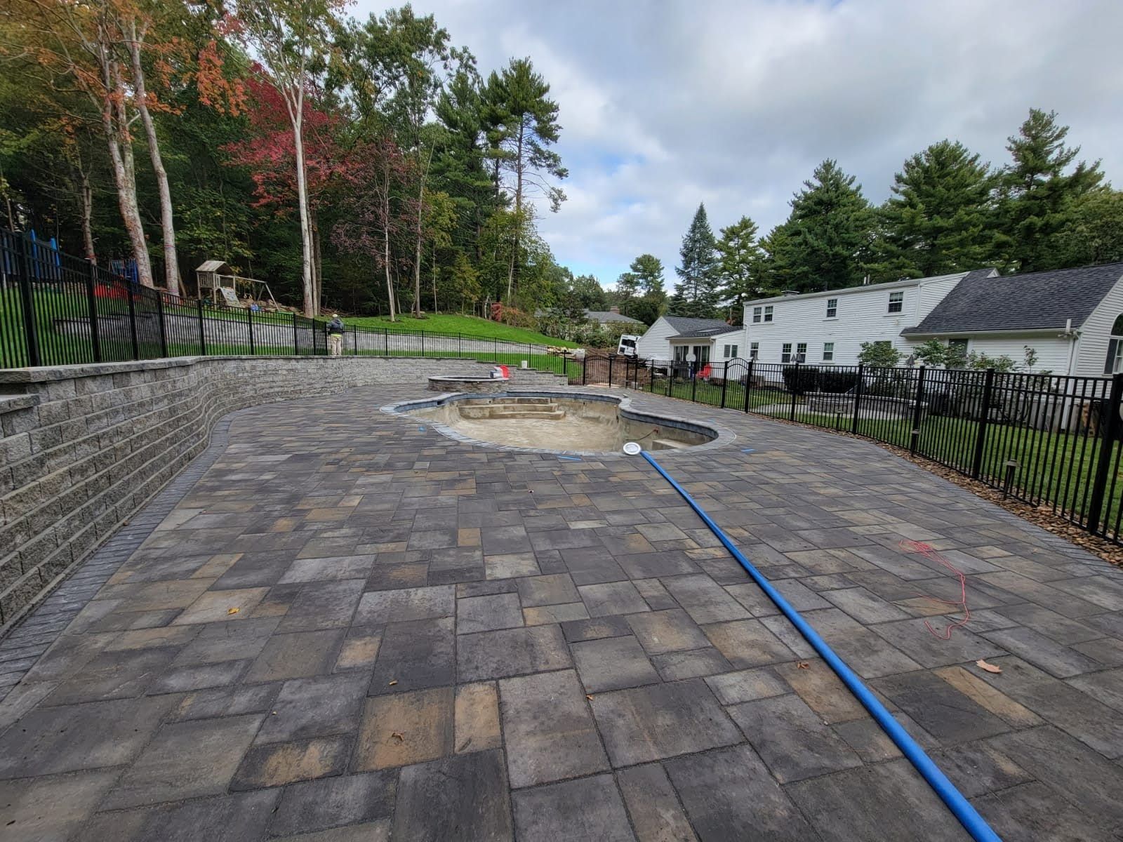 Stone-paver patio with a round fire pit, blue hose, and a fenced backyard beside houses and trees