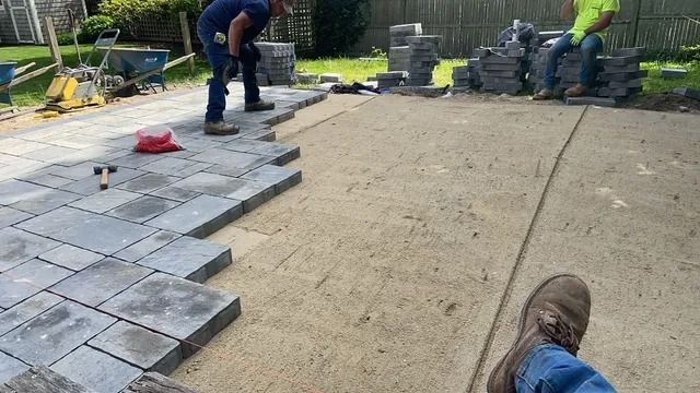 Workers laying gray paving stones on a patio beside stacked pavers and a concrete slab