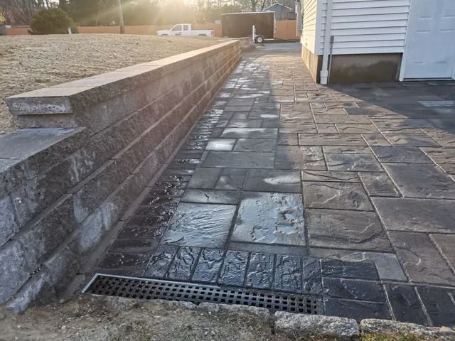 Stone patio walkway beside a retaining wall and house, with a drain grate in the foreground.