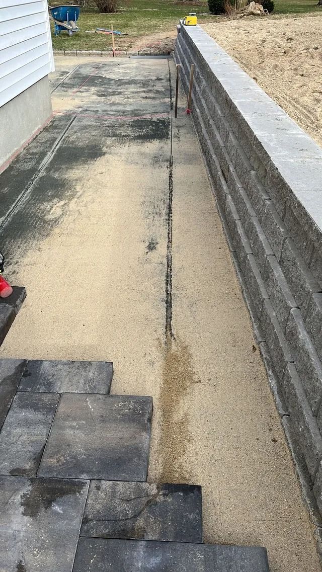 Narrow concrete walkway beside a house and retaining wall, with steps in the foreground and gravel above the wall