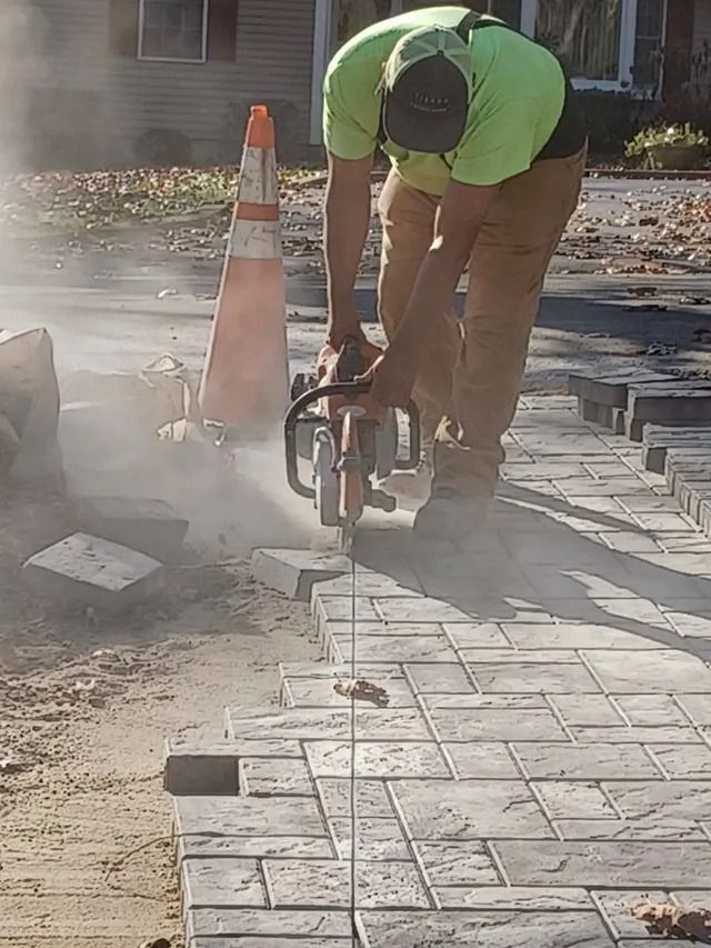 Worker cutting paving stones with a saw on a dusty sidewalk next to an orange traffic cone.