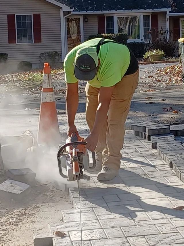 Worker cutting stone pavers with a saw on a driveway, with dust and traffic cones nearby