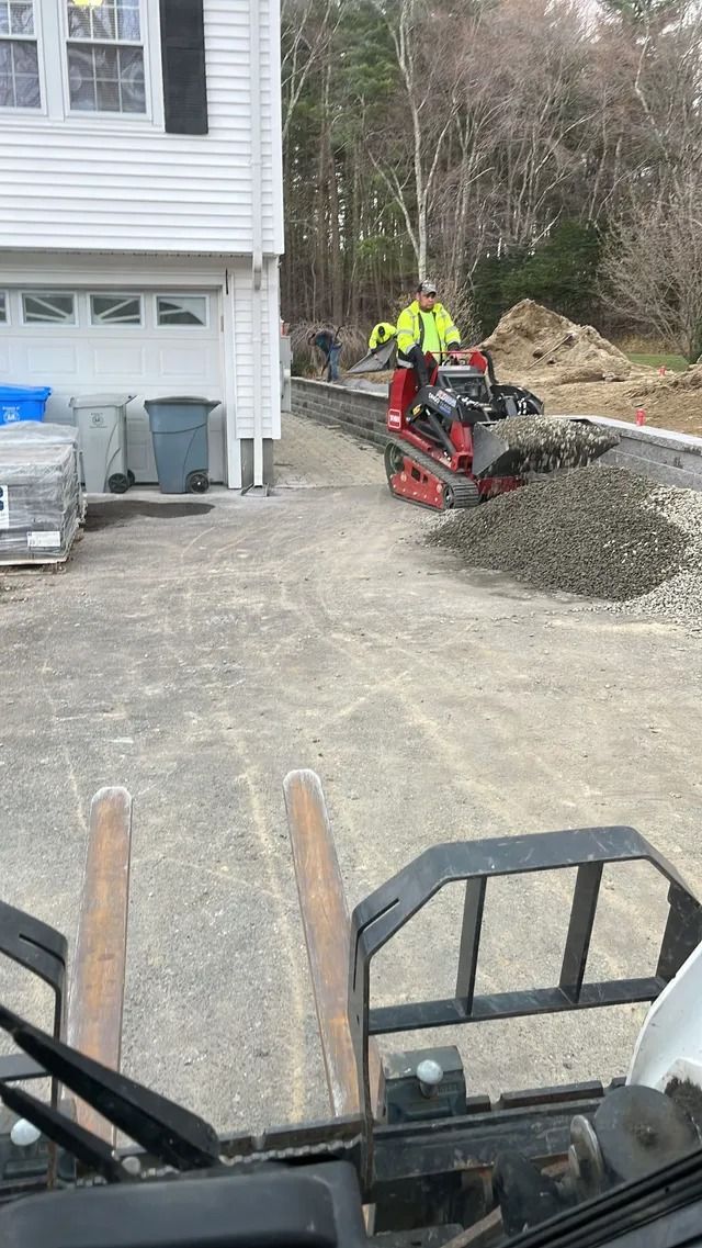 Worker in neon jacket operating a skid steer on a gravel driveway beside a house.