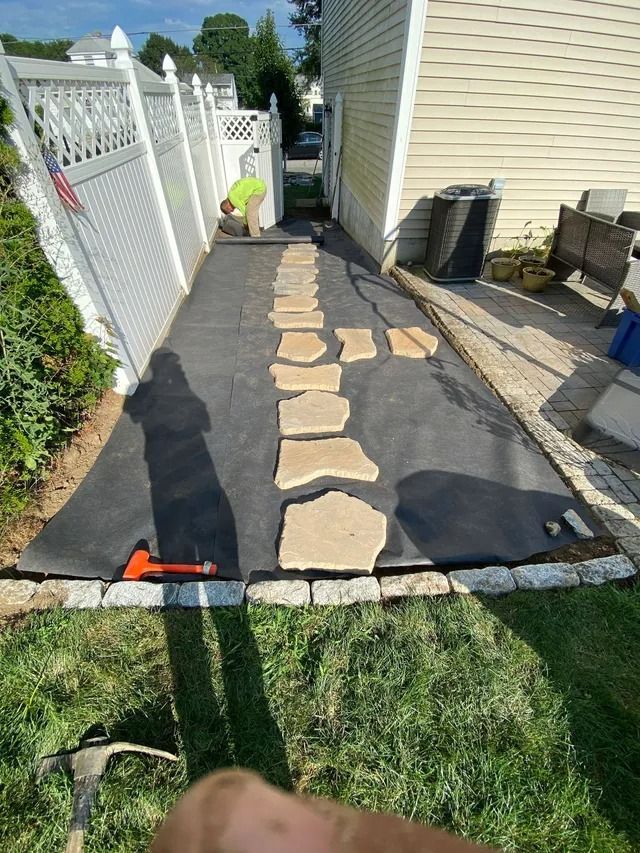 Narrow stone path under construction beside a house, bordered by black landscaping fabric and a white fence.