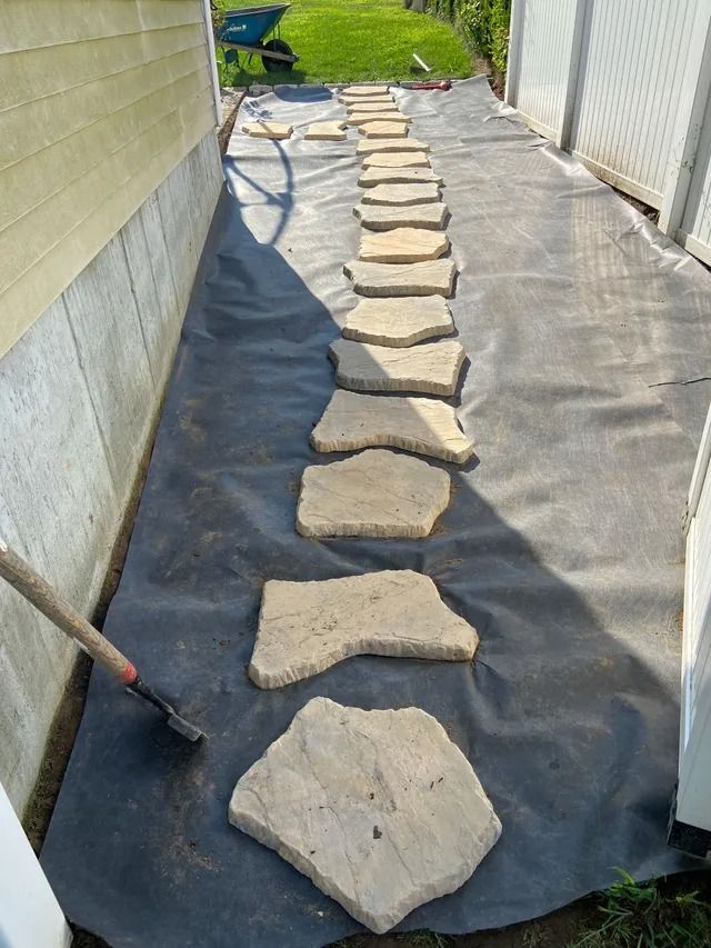 Stepping stones laid in black landscape fabric along a narrow outdoor walkway between fences.