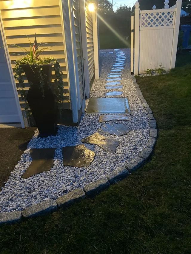 Curved stone walkway beside a house and white fence, lit by warm porch lights at night.