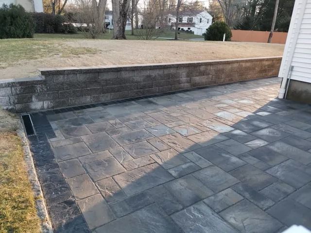 Paved patio beside a retaining wall and grassy yard, with a house in the background.