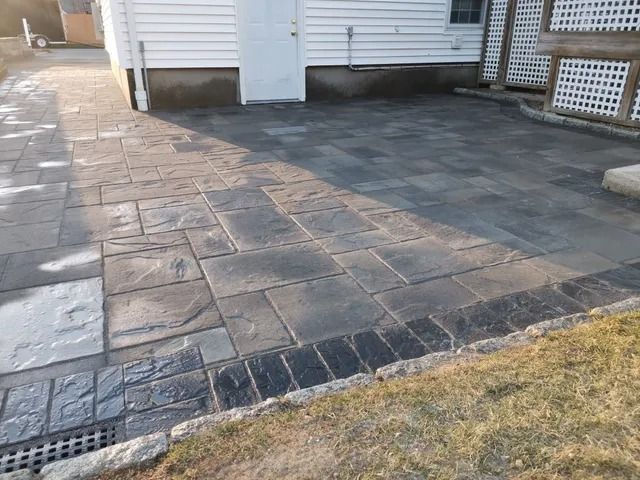 Paved driveway beside a white house with dark stone patio and a strip of grass in front