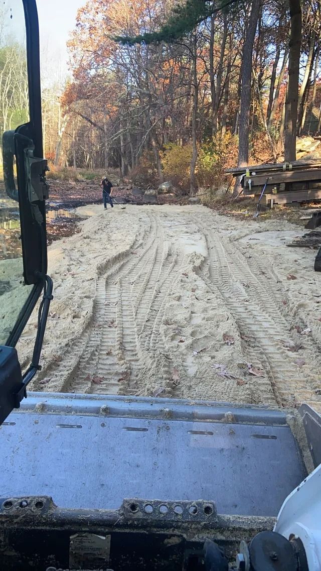 Bulldozer cab view of a sandy dirt road through autumn trees with stacked logs on the right
