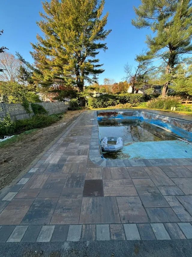 Stone patio beside a shallow blue pool, with tall evergreen trees and a garden in the background.