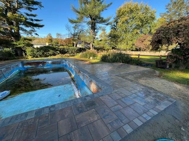 Backyard patio with an empty blue swimming pool, stone paving, and trees in the background