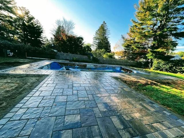 Stone patio beside a swimming pool and grassy yard under bright blue sky, with trees in the background