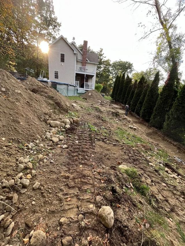 Steep dirt slope with rocks leading up to a white house at sunset, lined by tall evergreen trees