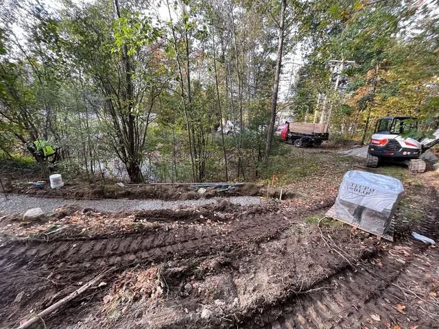 Wooded muddy lot with tire tracks, fallen logs, and parked cars near a stone marker