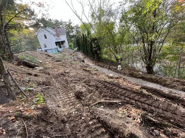 Rutted muddy hillside with a white house in the background and a person in a yellow vest near the river