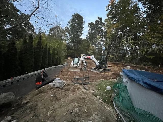 Construction site with excavator, dirt piles, and a partially built pool beside trees and fencing