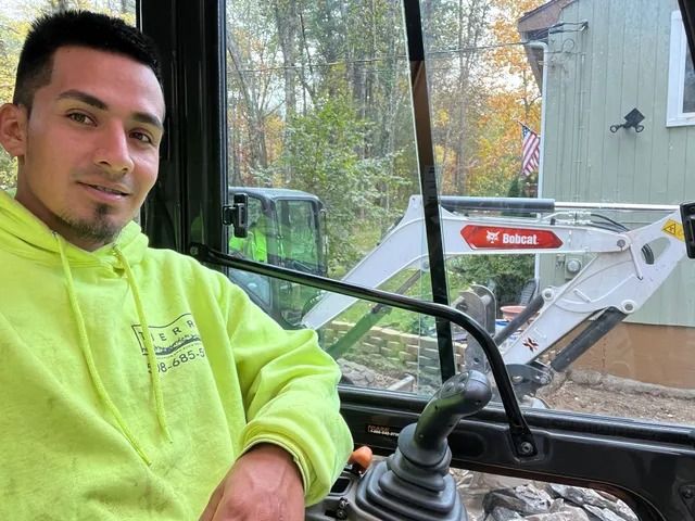 Person in a neon yellow hoodie sits in a vehicle cab with a white Bobcat loader visible outside the window.